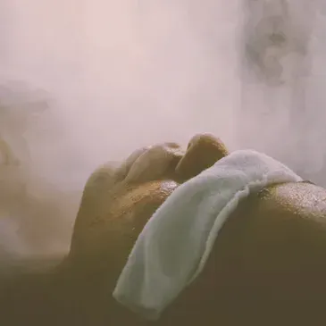 Close-up of a resting person’s arm with a white cloth draped over it in a hazy, steam-filled setting