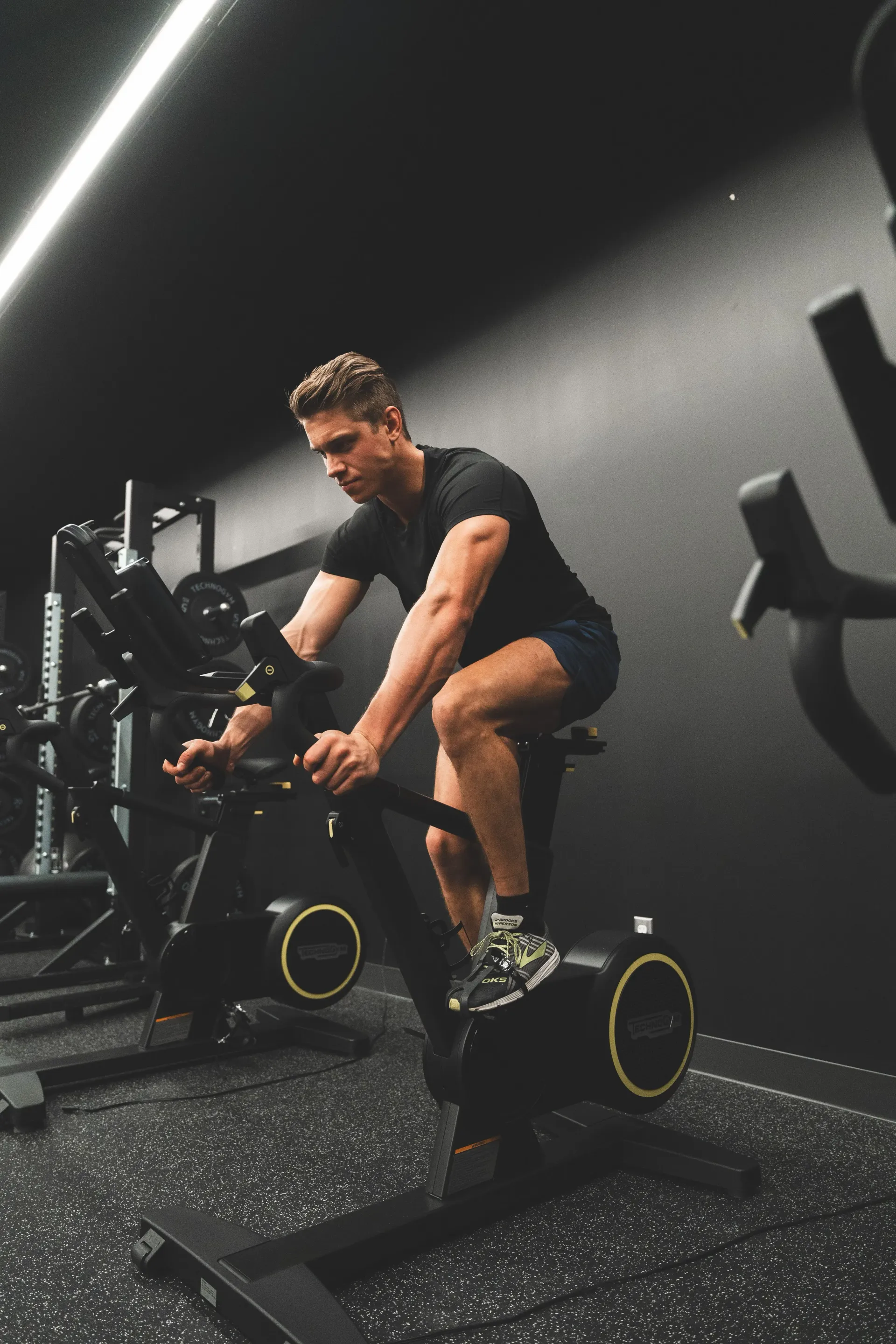 Man riding a stationary bike in a dim gym, wearing black workout clothes and focused on exercise