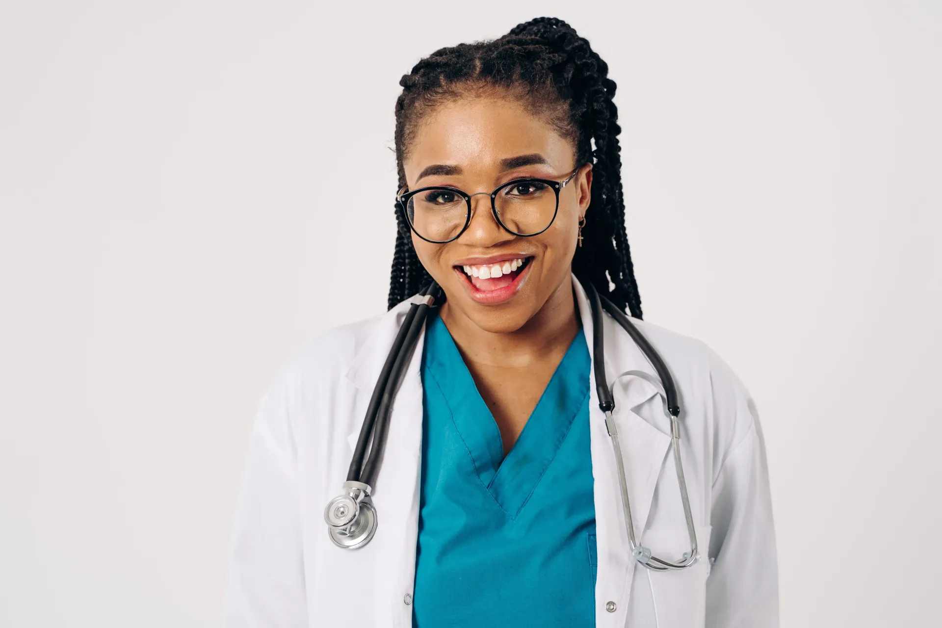 Smiling doctor in white coat and teal scrubs with stethoscope against a plain background