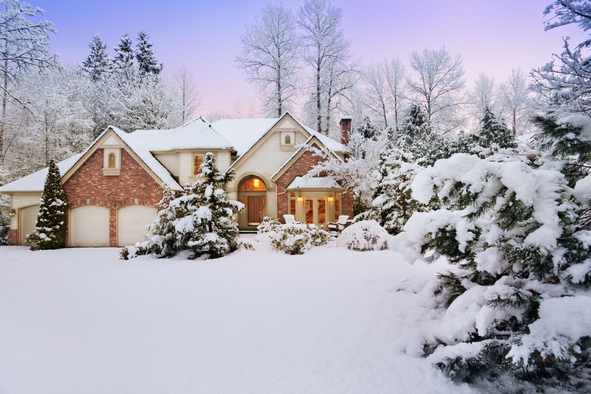 Snow-covered brick house with lit windows and evergreen trees in a winter landscape