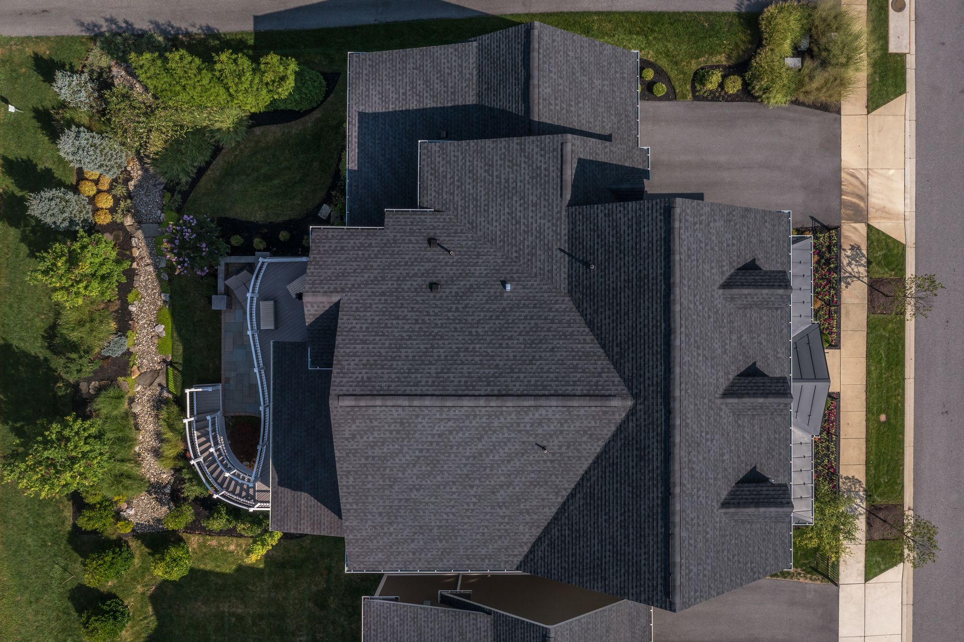 Aerial view of a dark-roofed house with a driveway, lawn, and trees.