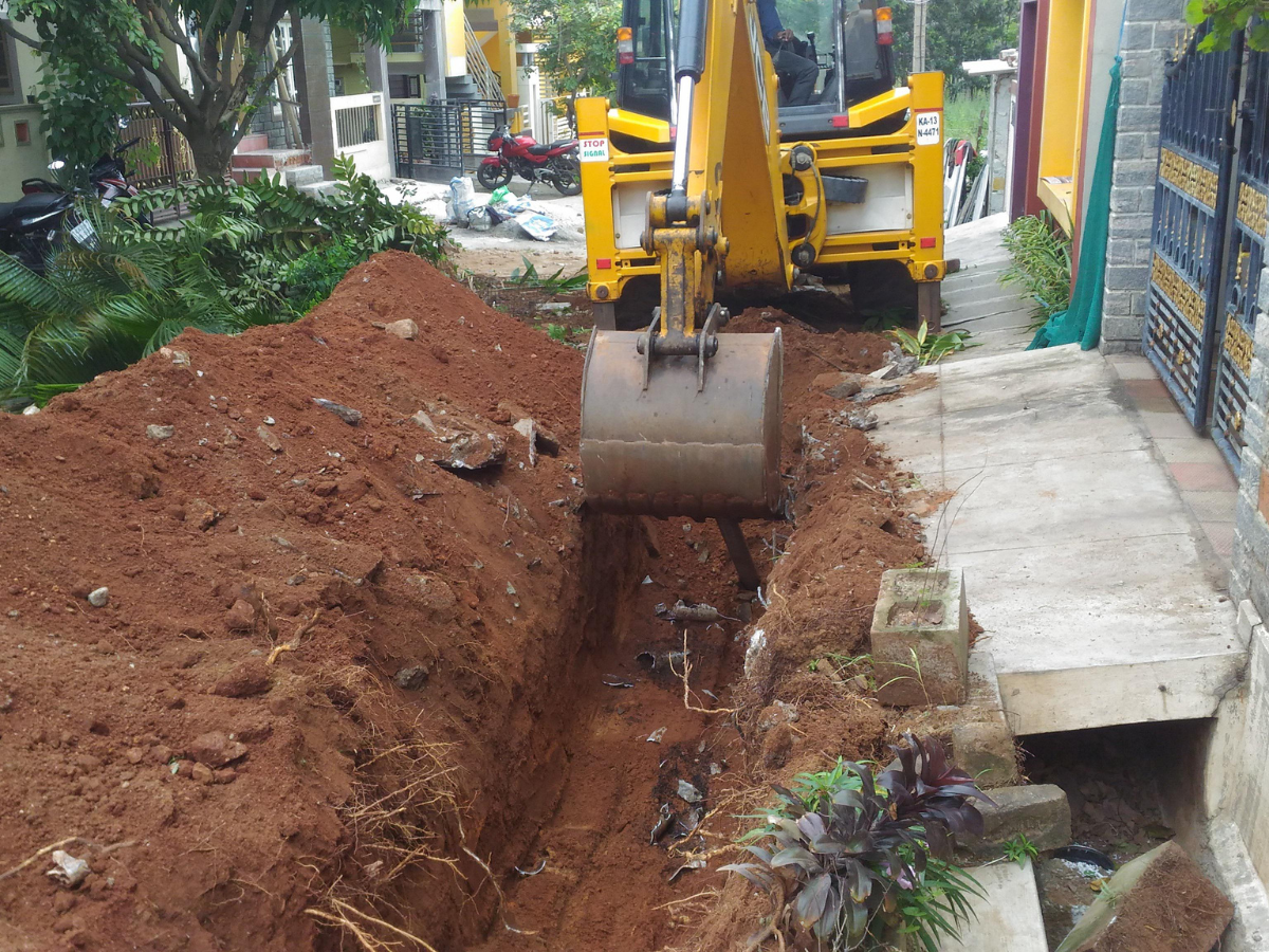Yellow excavator digging a trench beside a sidewalk, exposing pipes.
