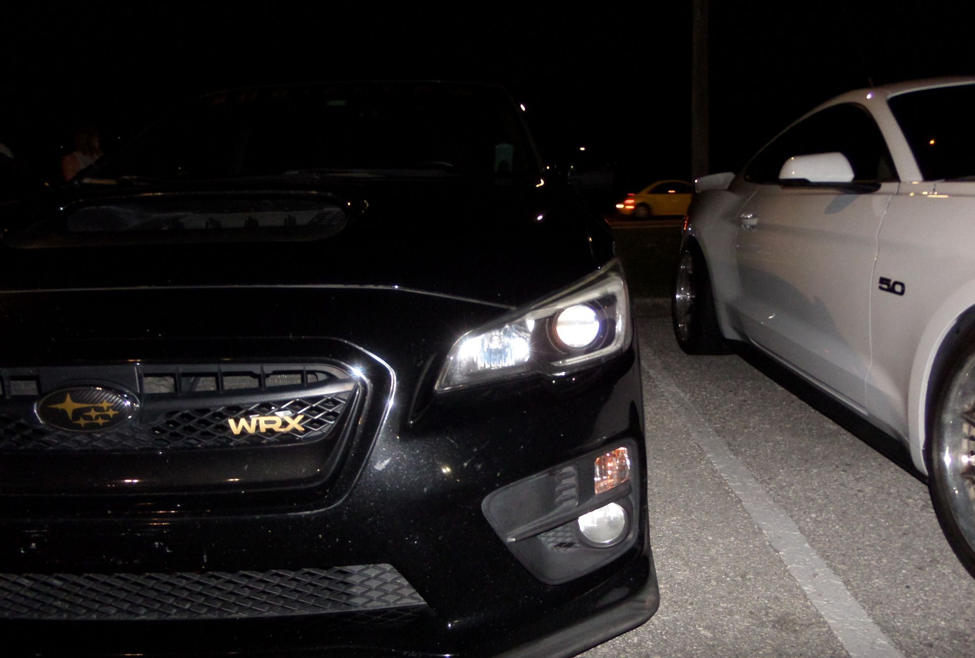 A black subaru wrx is parked next to a white mustang in a parking lot at night.
