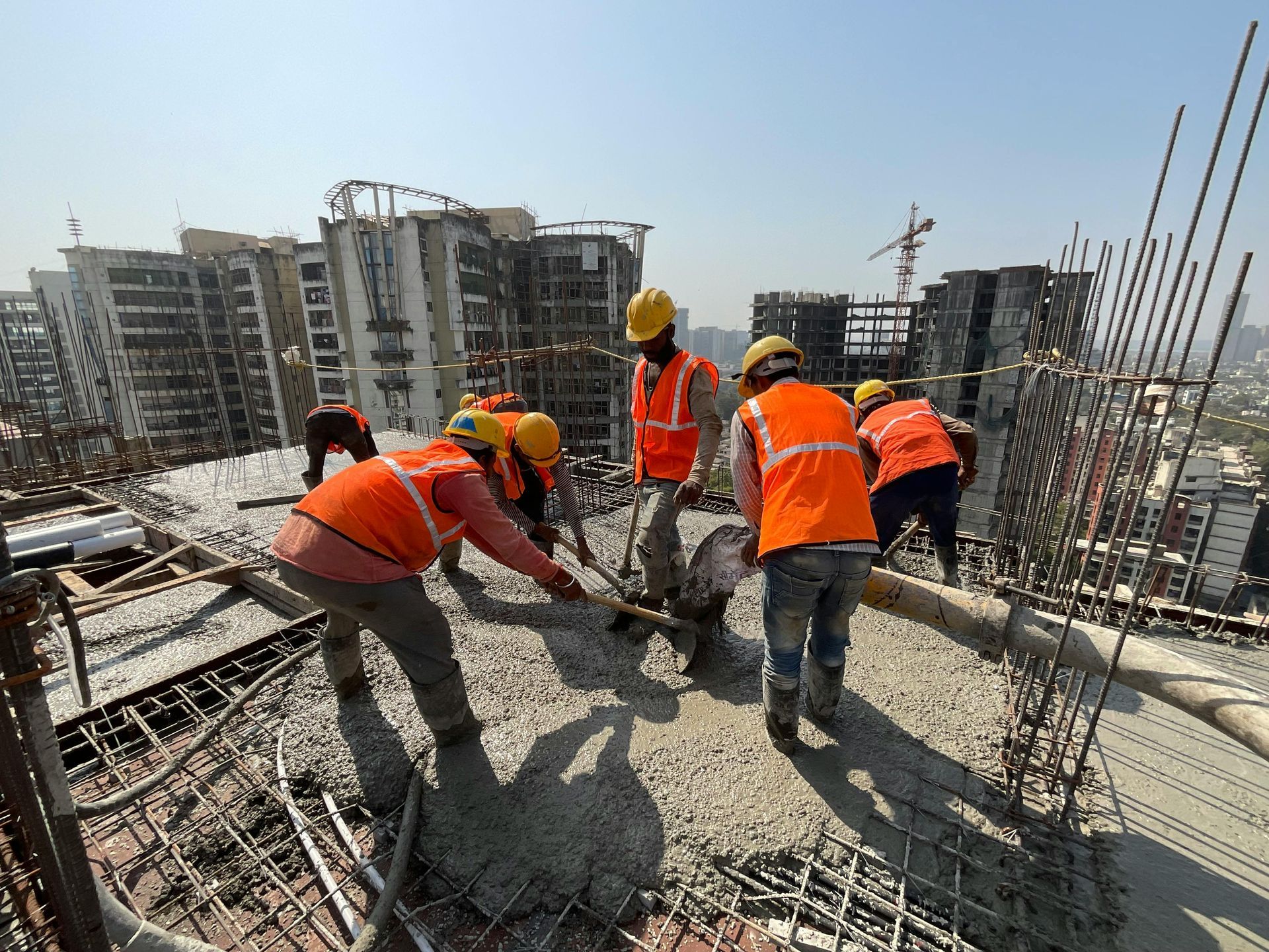 Construction workers in orange vests and hard hats pour concrete on a high-rise building rooftop.