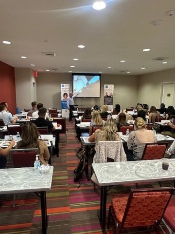 Conference room presentation with seated audience facing a speaker at the front of the room.