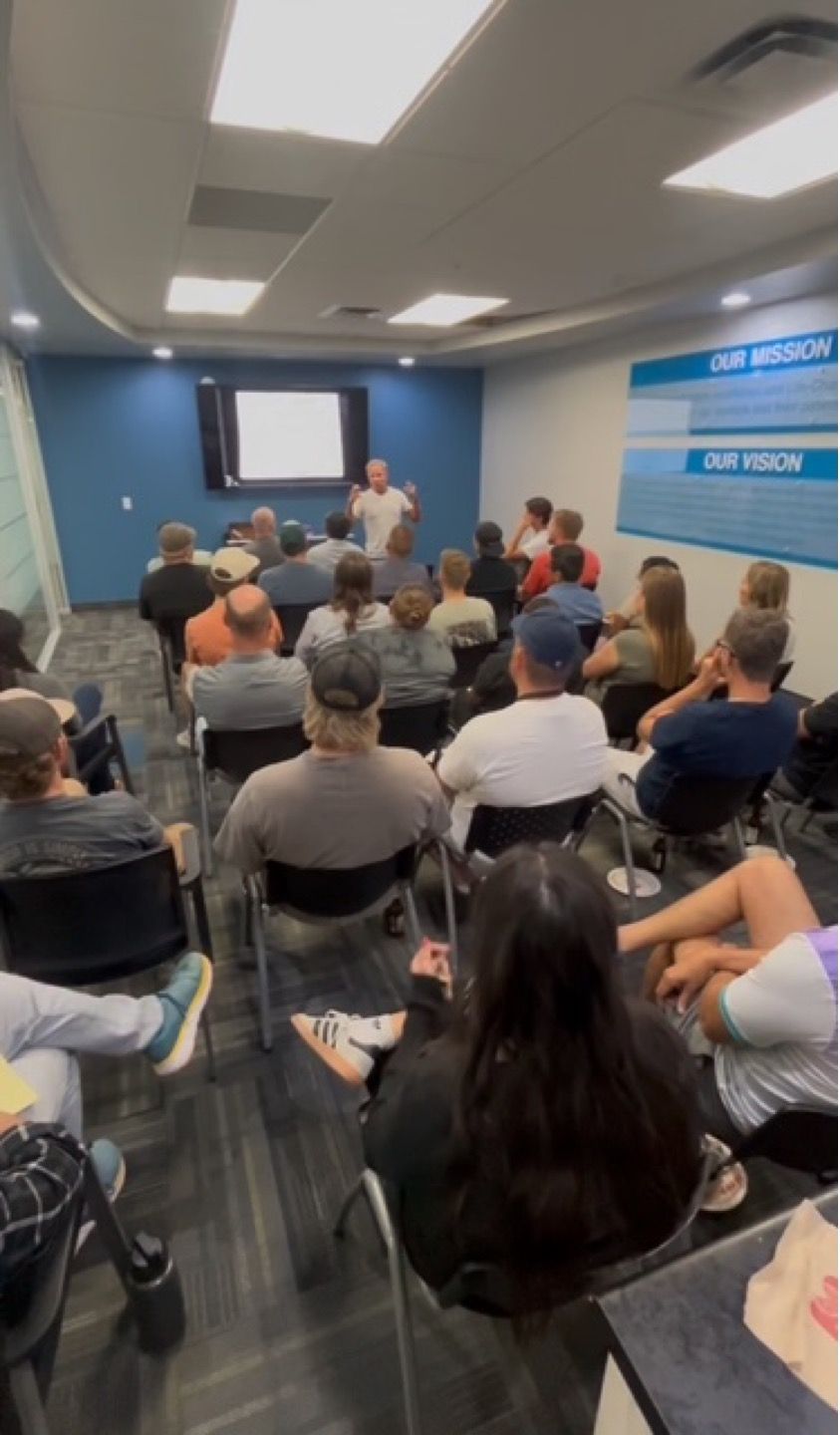 Meeting room audience facing a presenter near a screen in a blue-walled classroom, seated on folding chairs