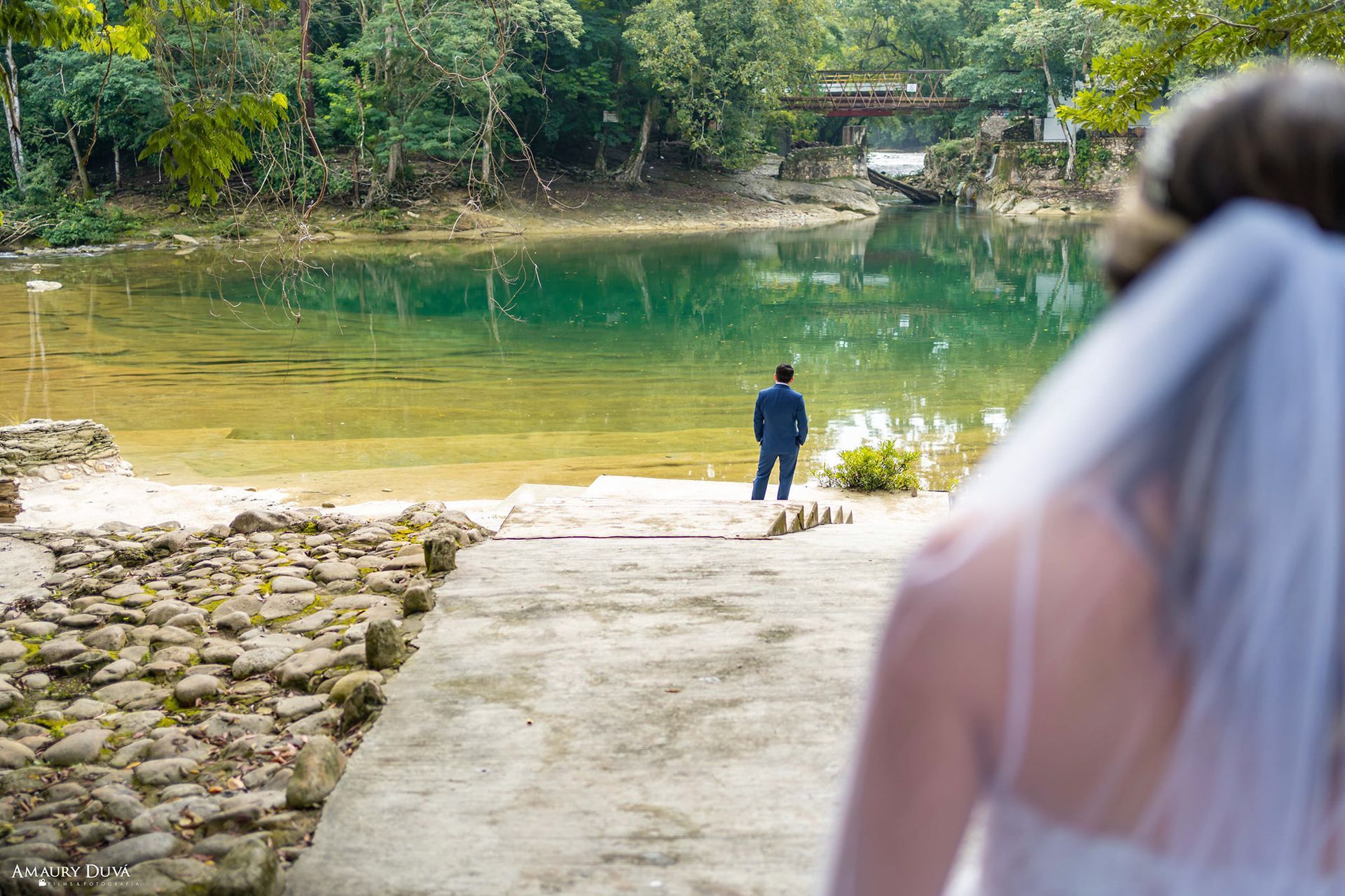 Una novia y un novio están de pie en un muelle mirando un lago.