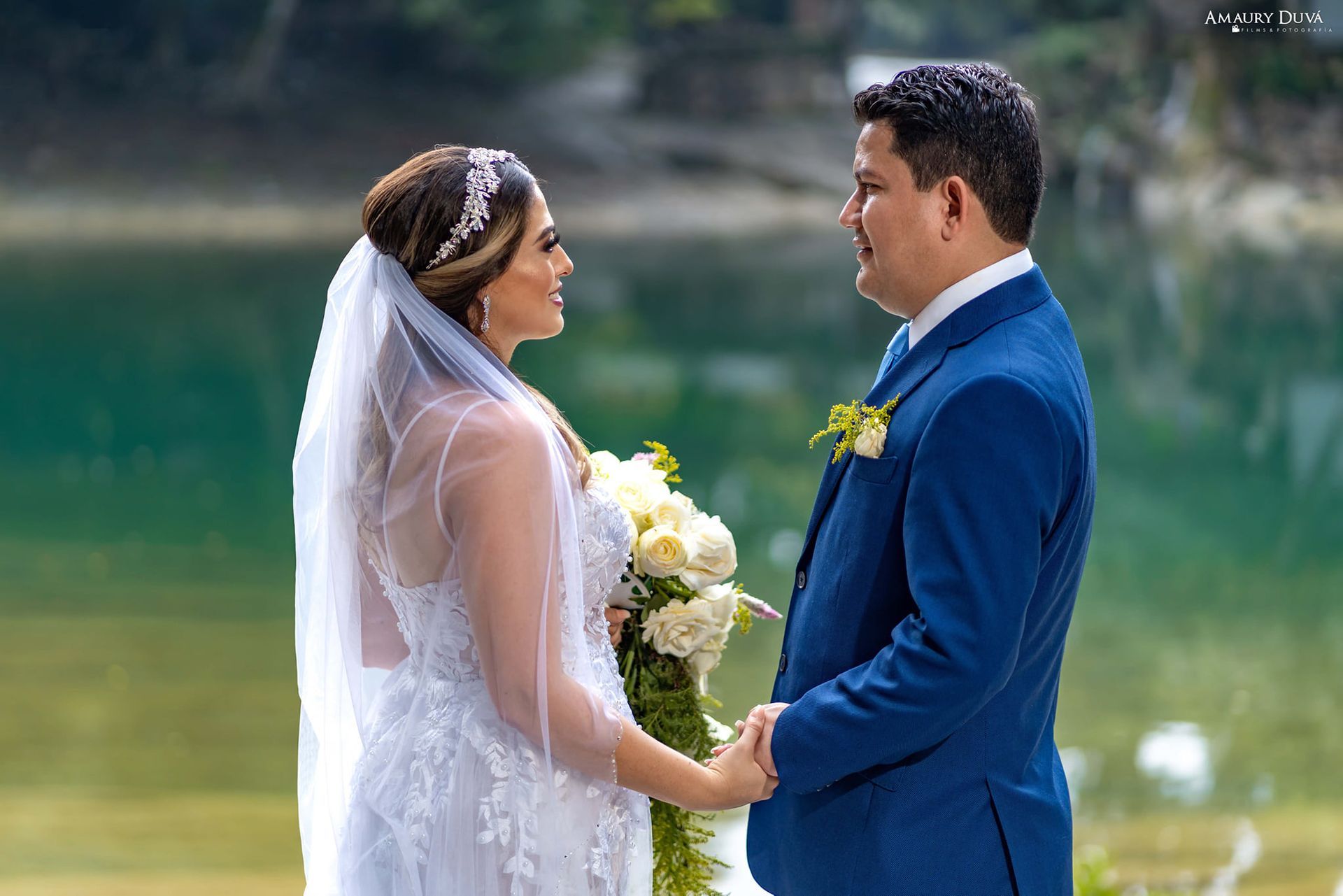 Una novia y un novio se toman de la mano y se miran frente a un lago.