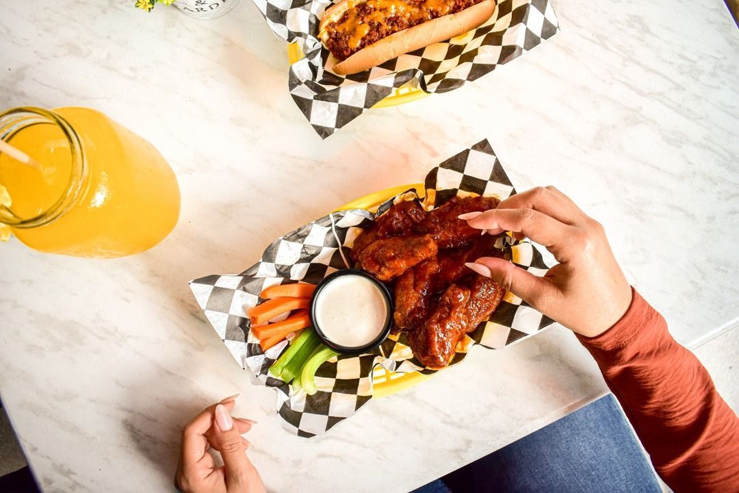 Una persona está sentada en una mesa comiendo alitas de pollo y un hot dog.