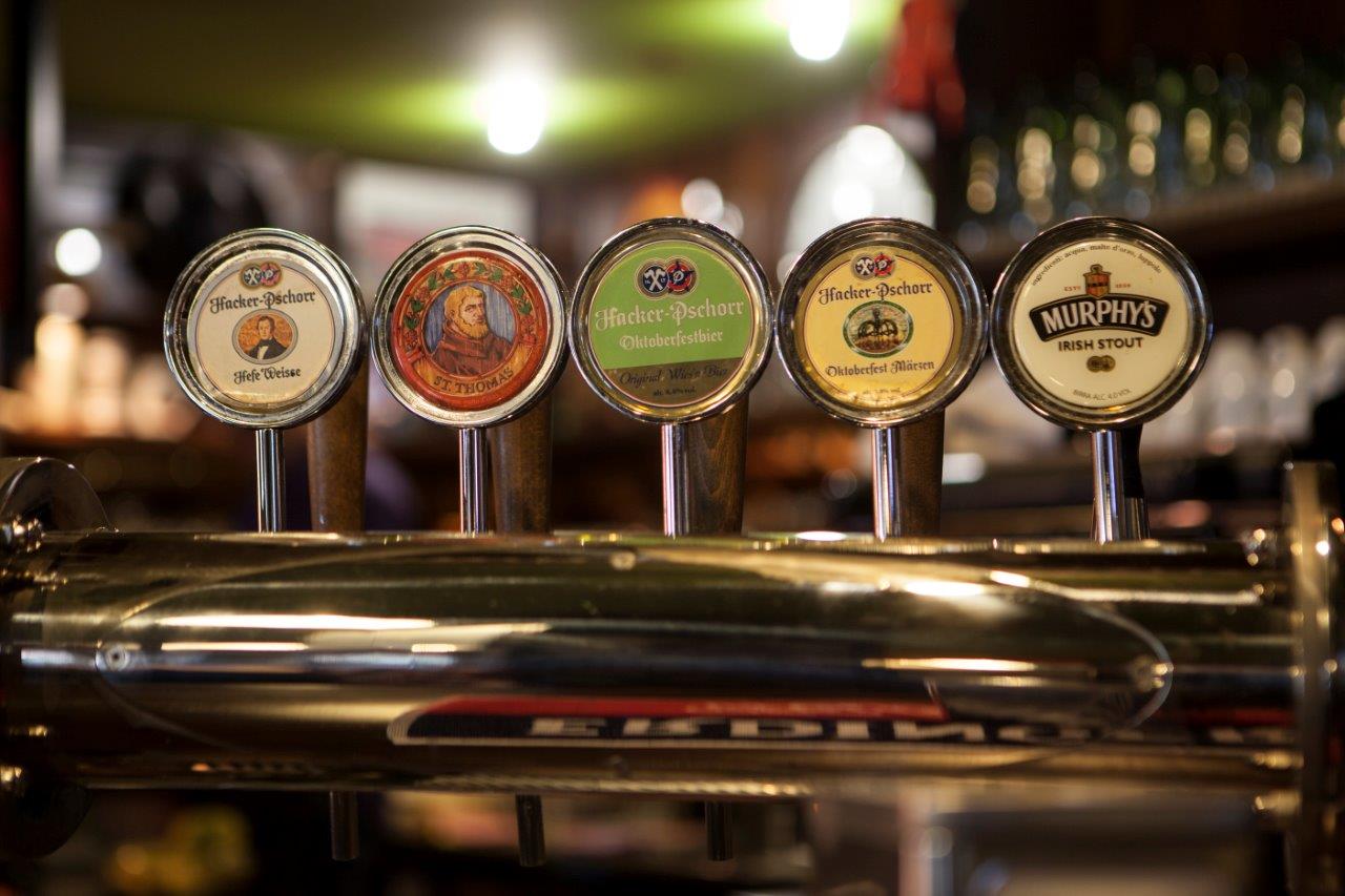 Cinque spine di birra con etichette di marca in un ambiente da bar.