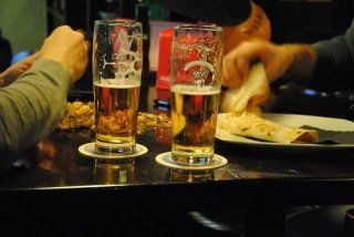 Due bicchieri di birra parzialmente vuoti sui sottobicchieri, accanto a cibo e persone sedute a un tavolo.