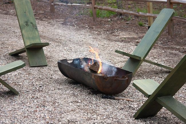 A fire pit is surrounded by wooden chairs in a gravel area.