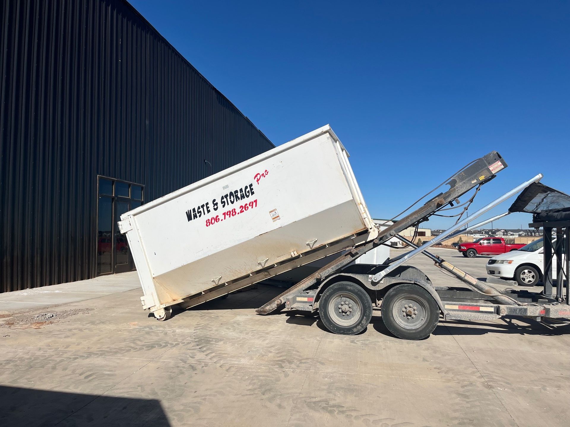 White dumpster tilted on a trailer, in front of a building on a sunny day.