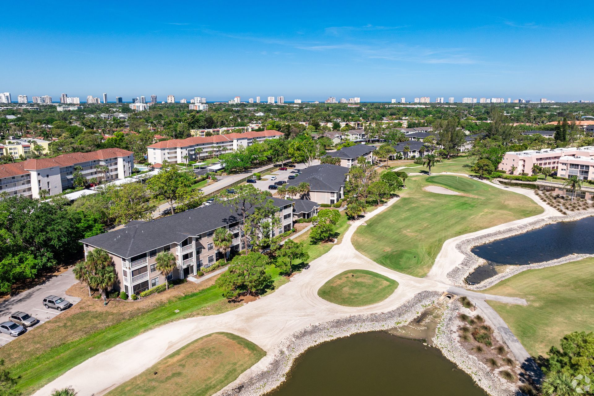 Aerial view of Belvedere at Quail Run and a golf course with winding paths, ponds, apartment buildings, and a distant city skyline