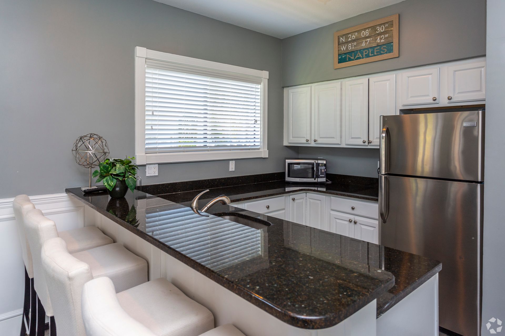 Modern clubhouse kitchen with white cabinets, granite island, stainless steel fridge, and barstools by a window