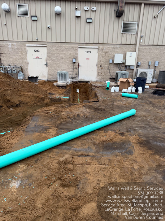Construction site with a large turquoise pipe in muddy ground beside a building wall.