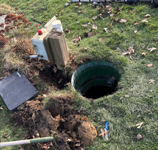 Open manhole with a yellow-and-black cable device and wires inside, surrounded by grass.