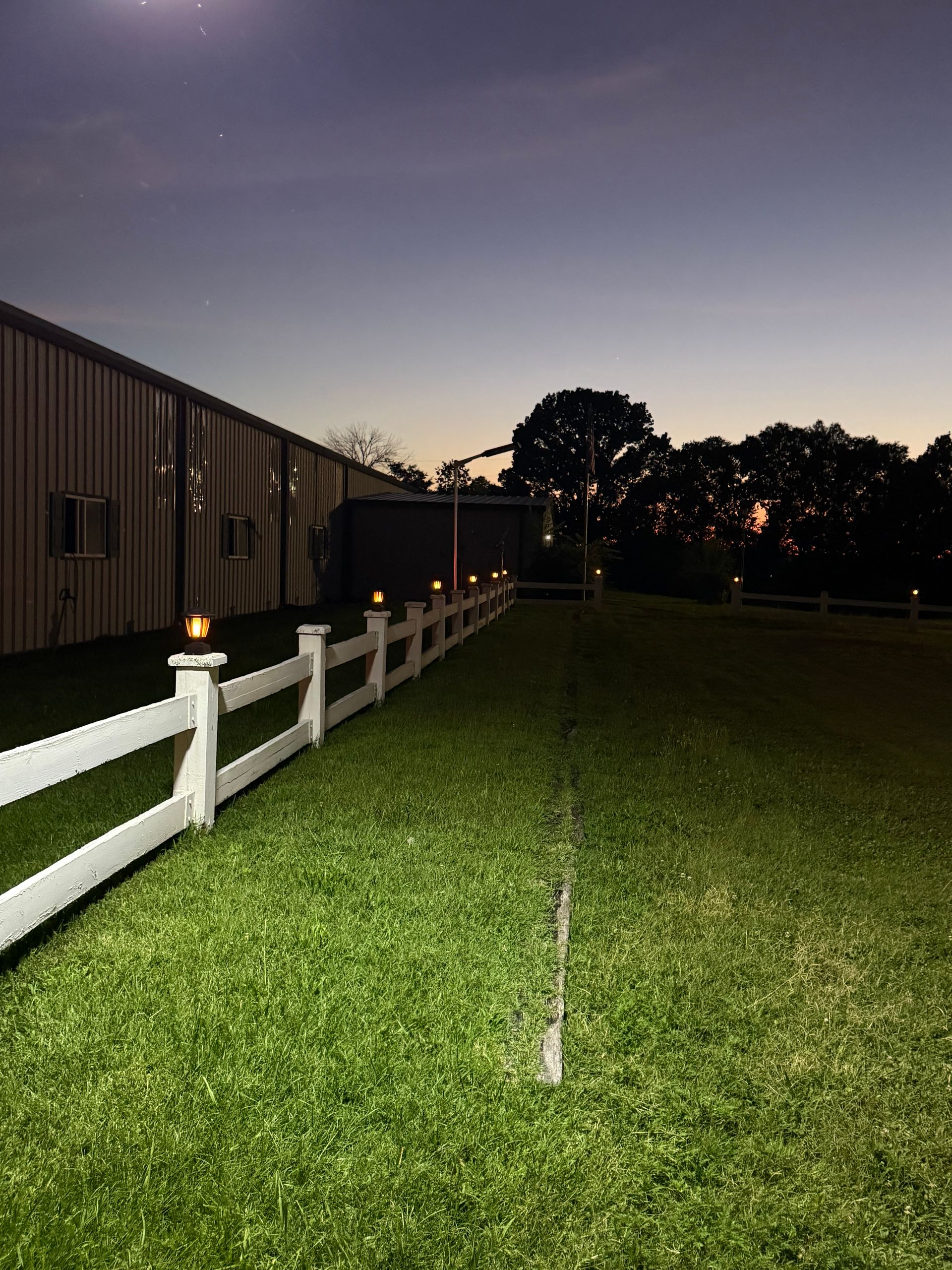 Nighttime grassy field beside a white fence and building, lit by a few small lights.