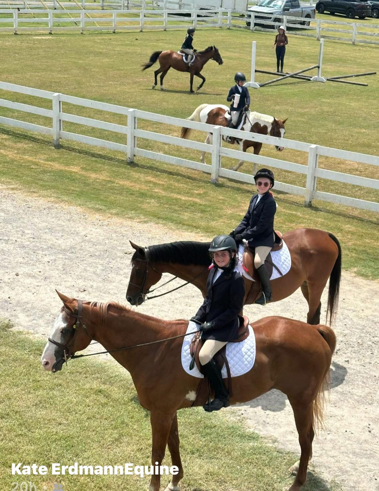 Two riders on brown horses in a grassy arena, with other riders and white fences in the background