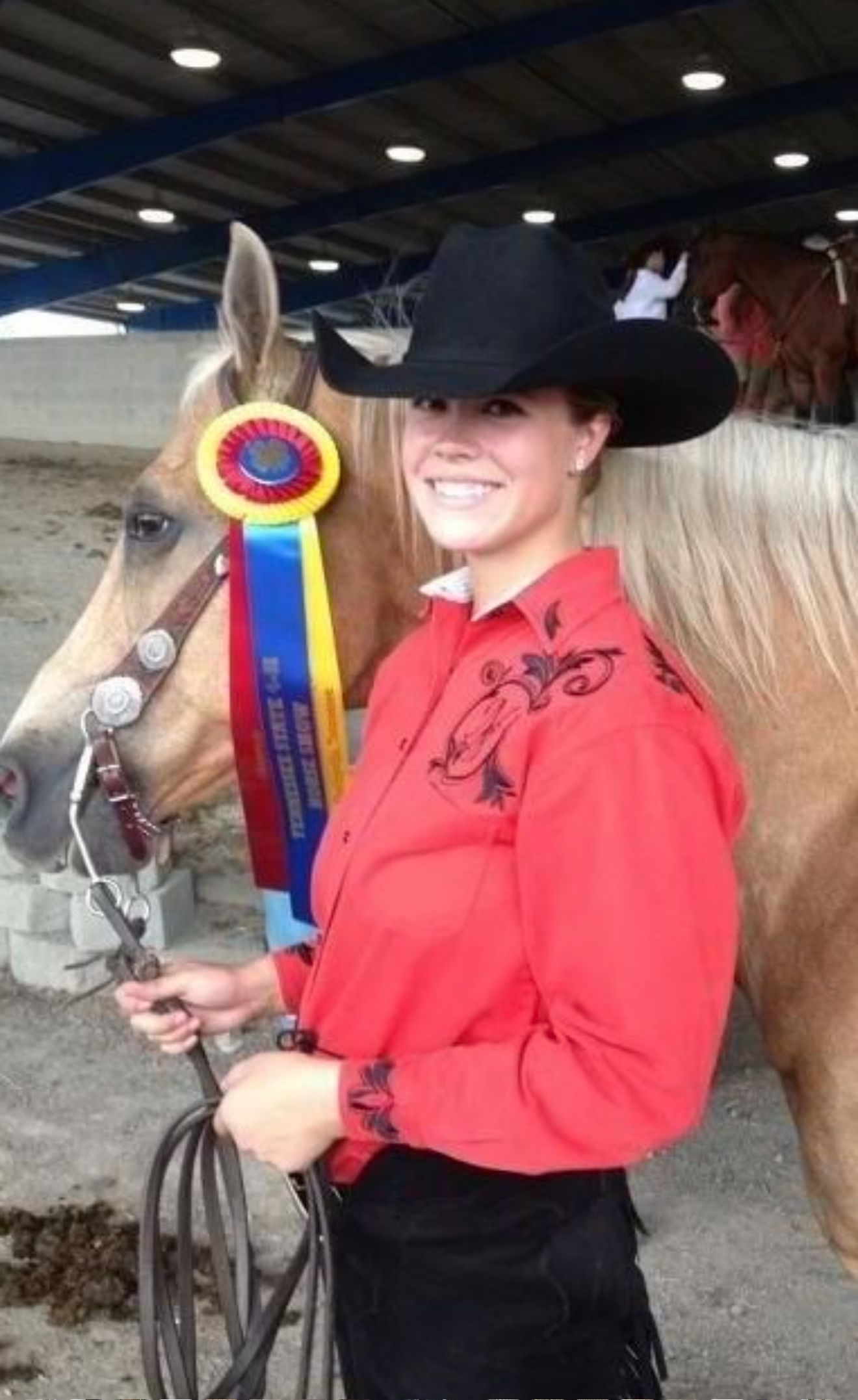 Woman in black cowboy hat holding a horse with a blue-and-yellow ribbon at an indoor arena