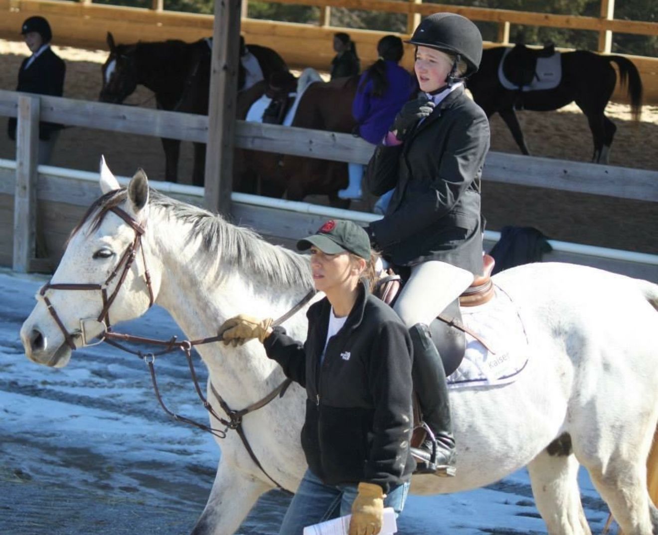Person riding a white horse in an outdoor arena, with another rider and horses in the background