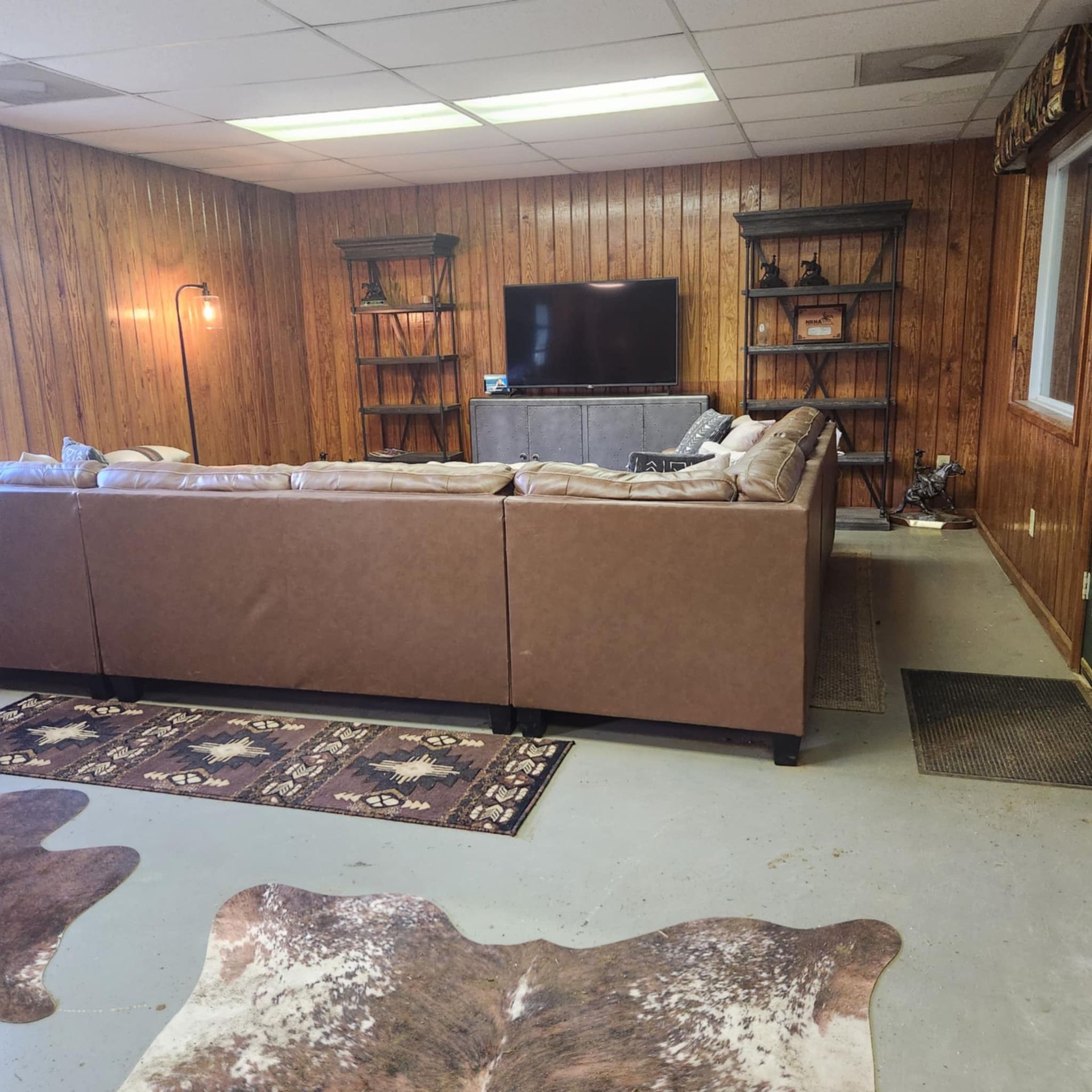 Basement living room with brown sectional, TV, wood-paneled walls, rugs, and shelves with decor