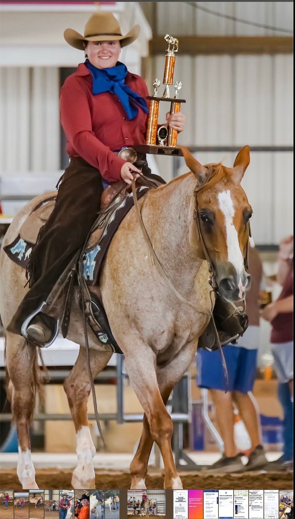 Cowgirl on a tan horse holds a trophy in an indoor arena.