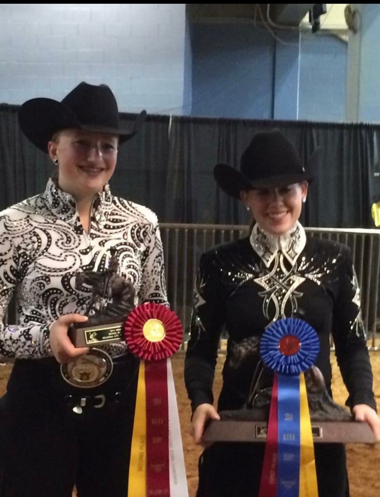 Two riders in black cowboy hats holding trophies with red and blue ribbons on a stage