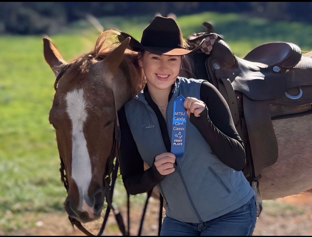 Woman in cowboy hat holding blue ribbon beside a horse with saddle in a sunny field