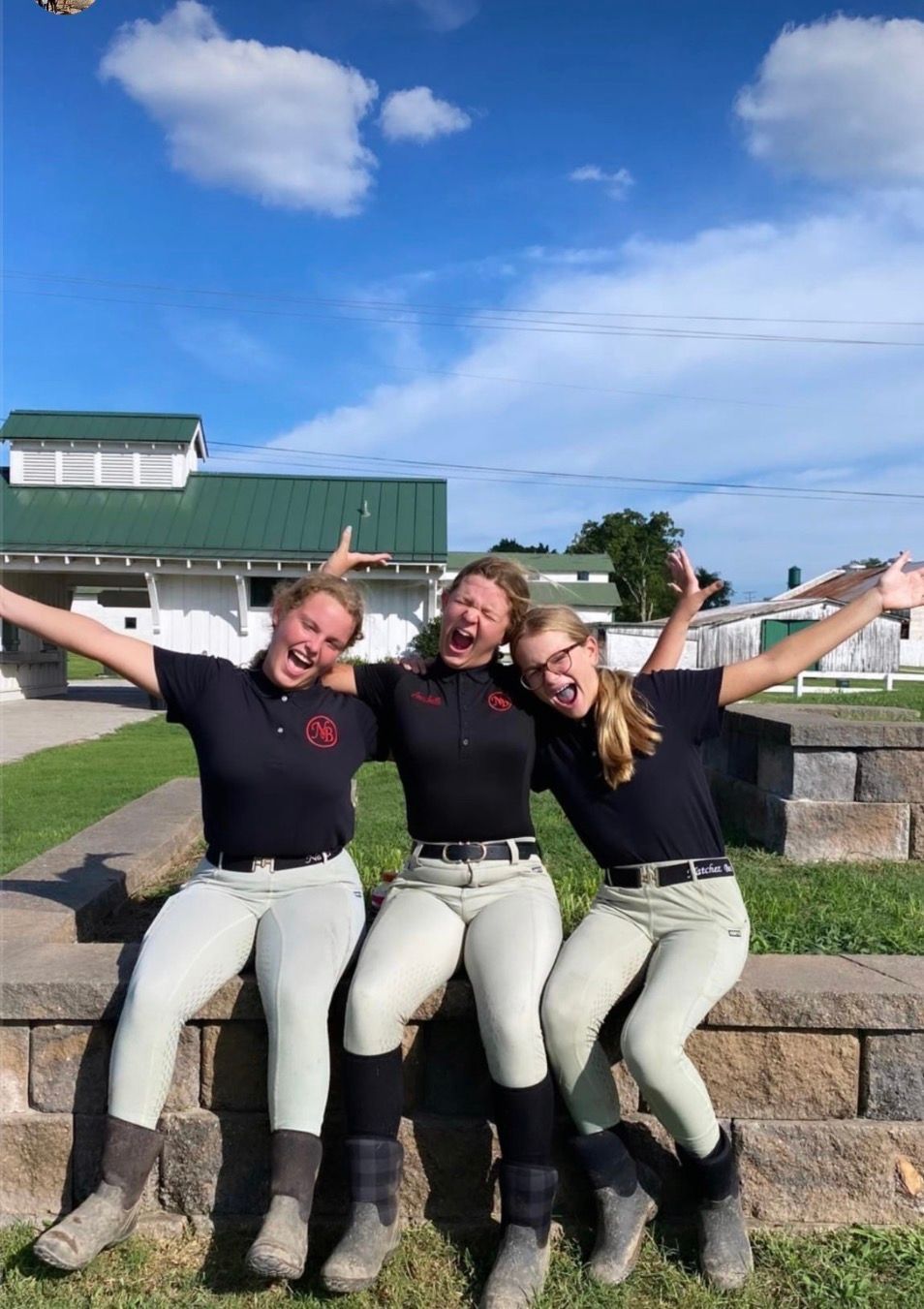 Three people in black shirts and light pants sitting on a stone wall, smiling with arms raised outdoors.