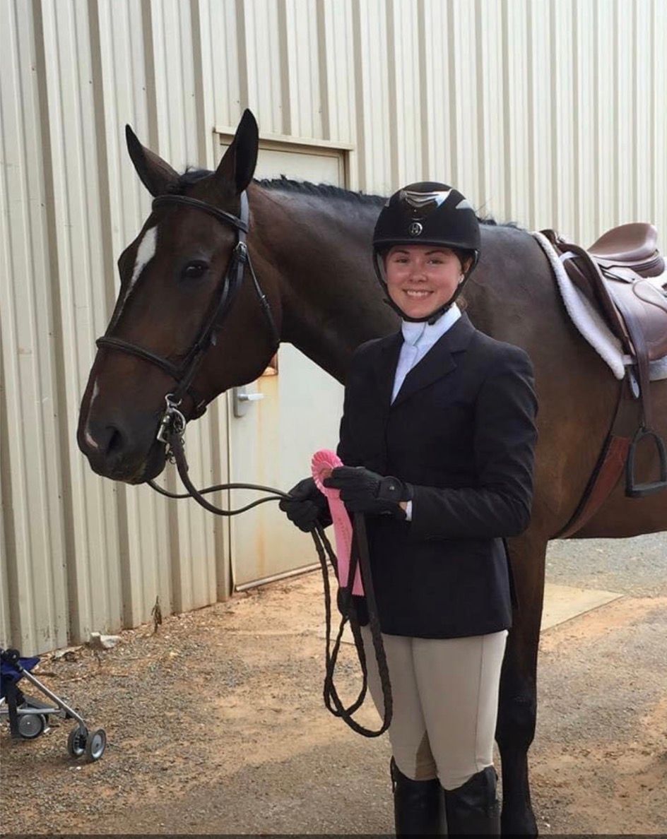 Rider in black show jacket and helmet stands beside a saddled brown horse in an outdoor stable area