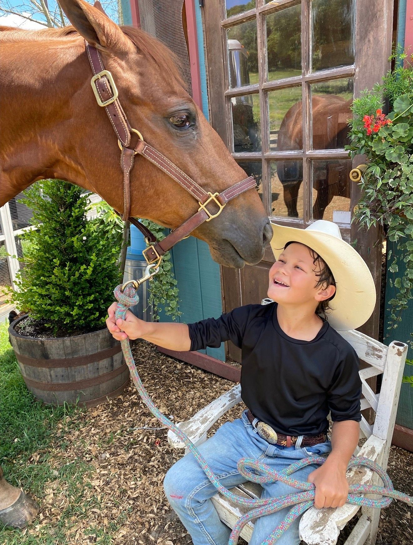 Child in a cowboy hat smiling at a horse, holding its lead rope outside a rustic building.