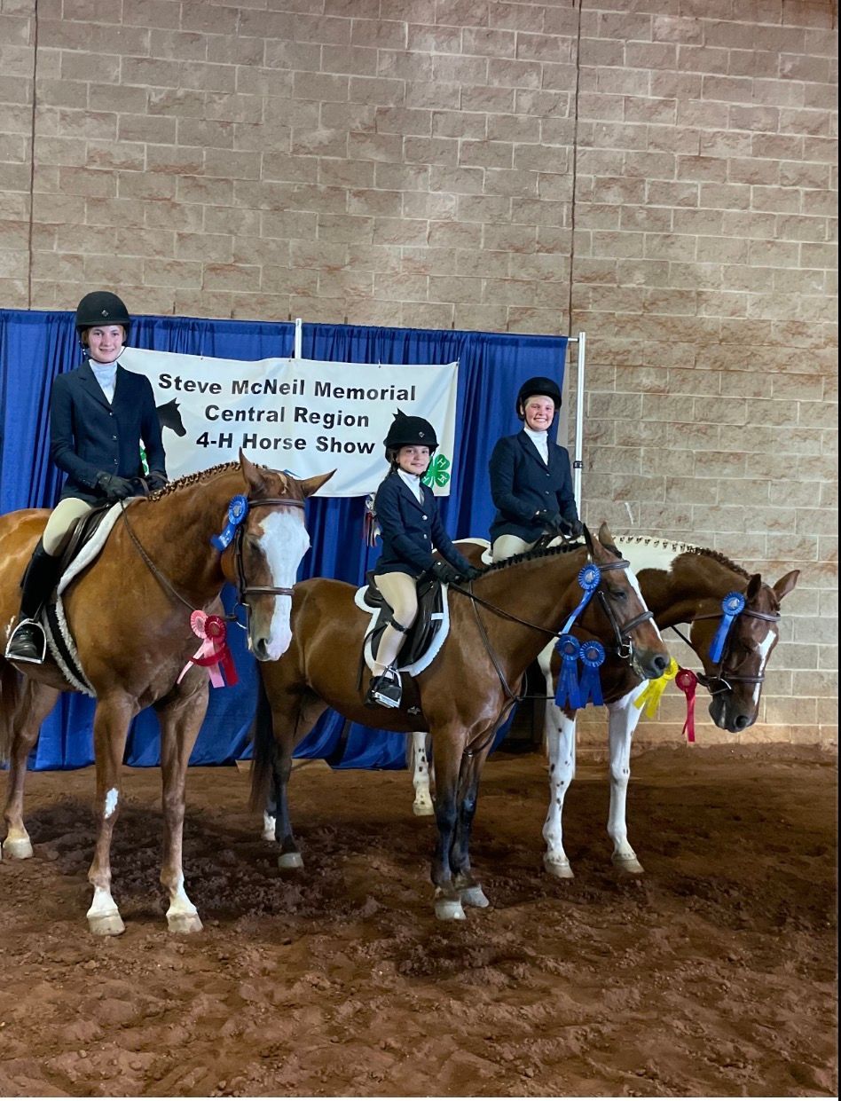 Three riders on horses pose indoors in front of a blue banner.