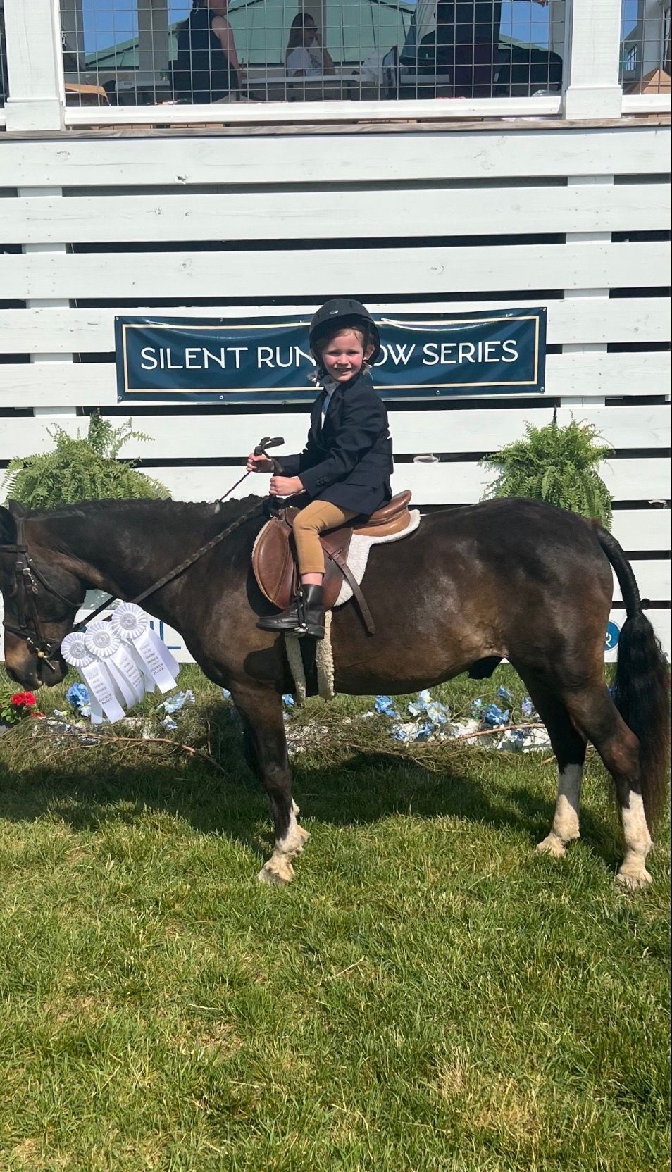 Person riding a dark brown horse on grass in front of a white building with a sign.