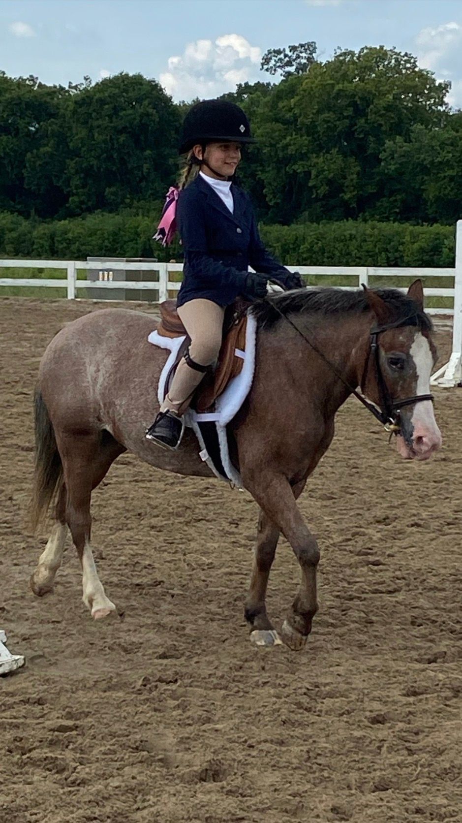 Child riding a brown pony in an outdoor arena, wearing a helmet and show attire.
