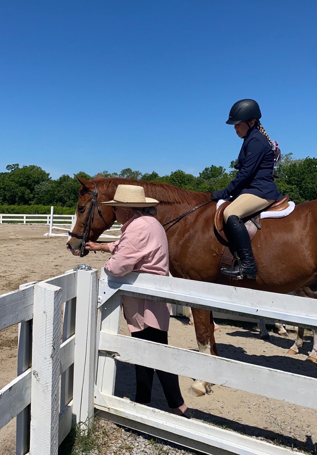 Person on a brown horse by a white fence, with a handler in a helmet nearby under a clear blue sky