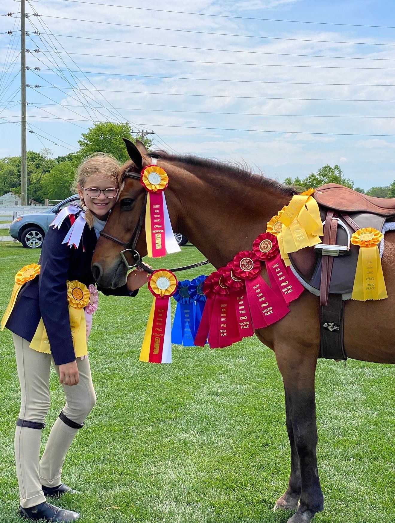 Girl posing beside a horse wearing award ribbons at a grassy outdoor show