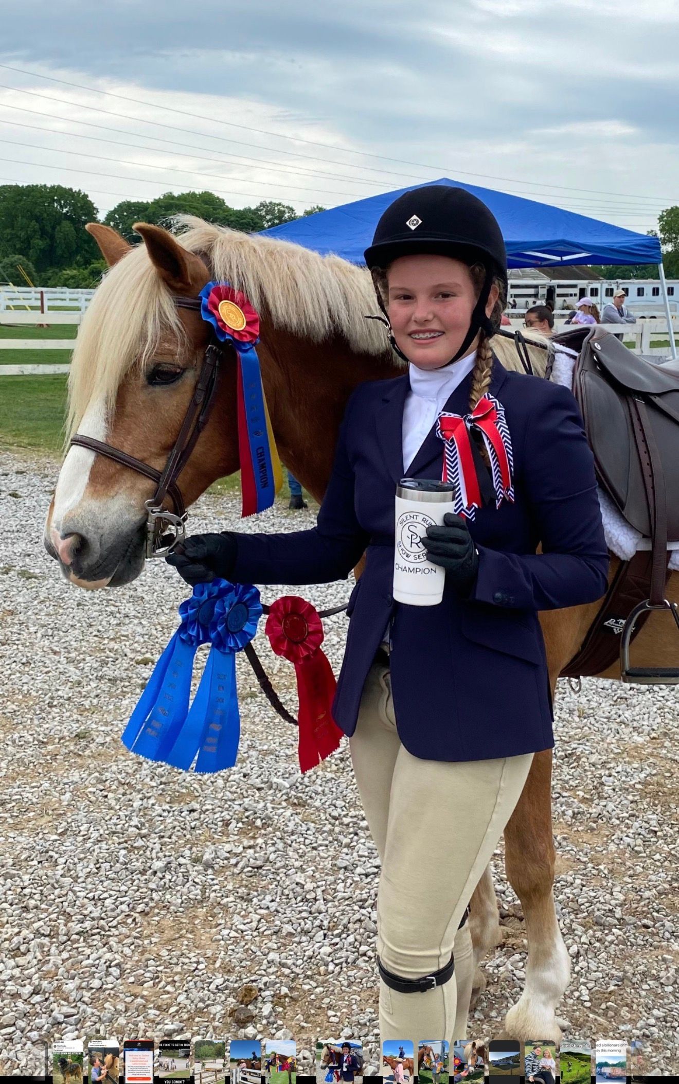 Smiling rider holding a tan horse with blue and red ribbons at an outdoor show.
