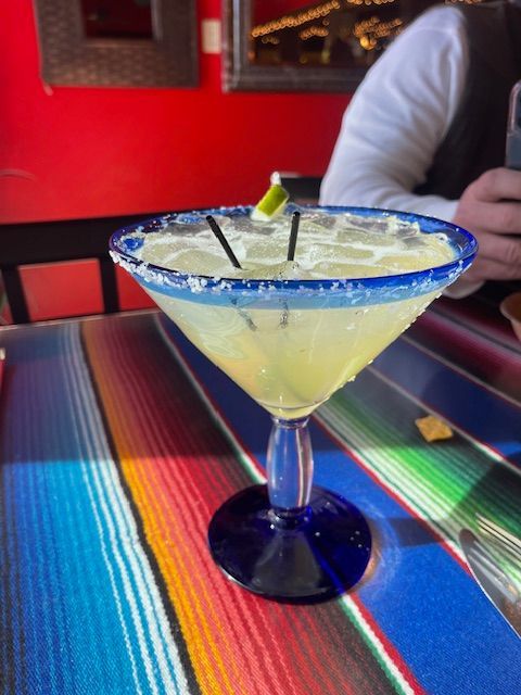 Margarita in a blue-rimmed glass, salt on the rim, served on a colorful striped table cloth.