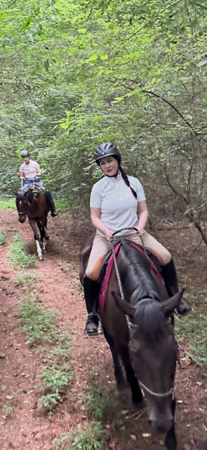 Two people riding horses on a dirt path in a wooded area. The lead rider smiles at the camera.