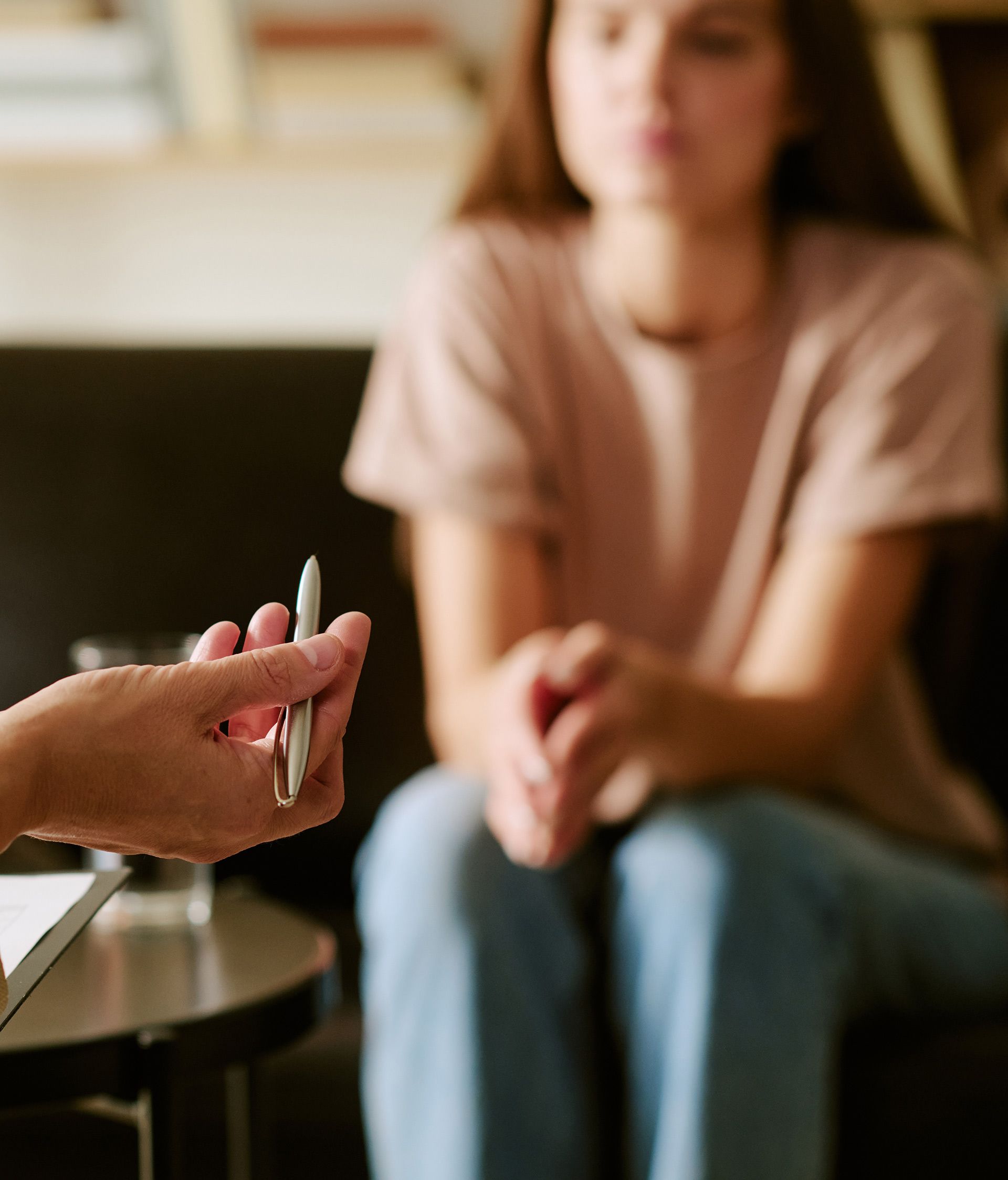 Therapist's hand holding pen, patient in background seated on a couch, focused.