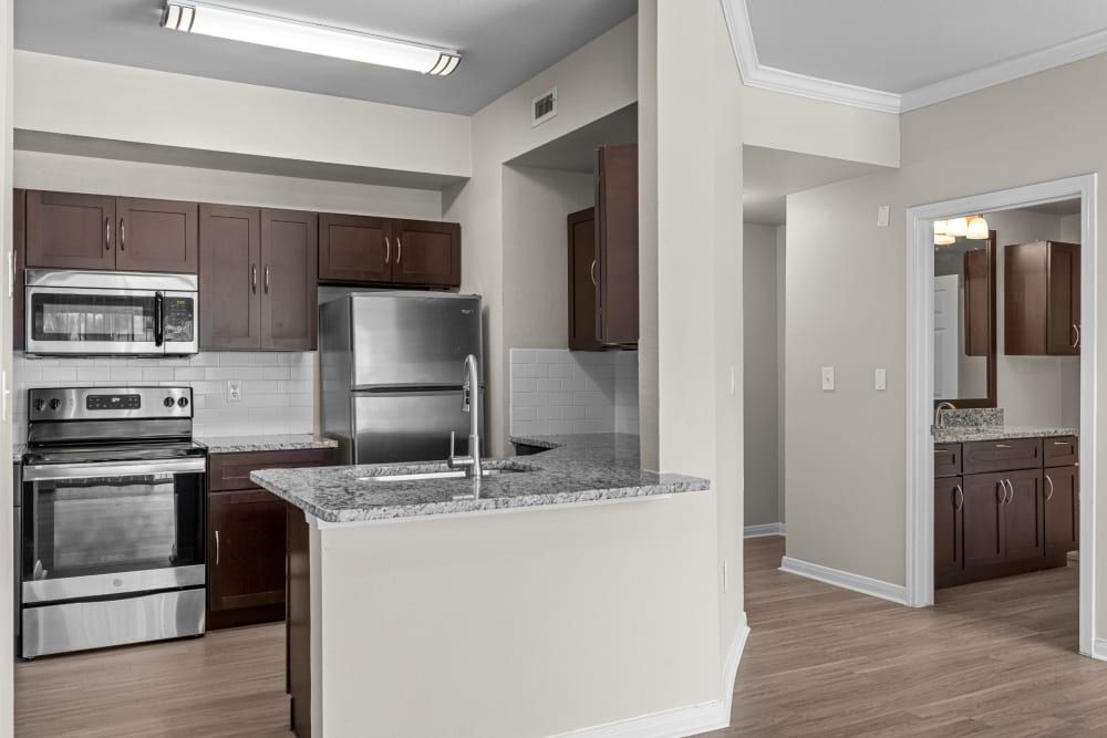 Modern kitchen interior with granite countertops and stainless steel appliances, including a refrigerator, microwave, and oven. The kitchen features dark brown wooden cabinets and light gray subway tile backsplash. Recessed and under-cabinet lighting illuminate the space, which has beige walls and a light wood laminate floor that extends into the adjoining room at Brooks on Preston in Plano, TX.