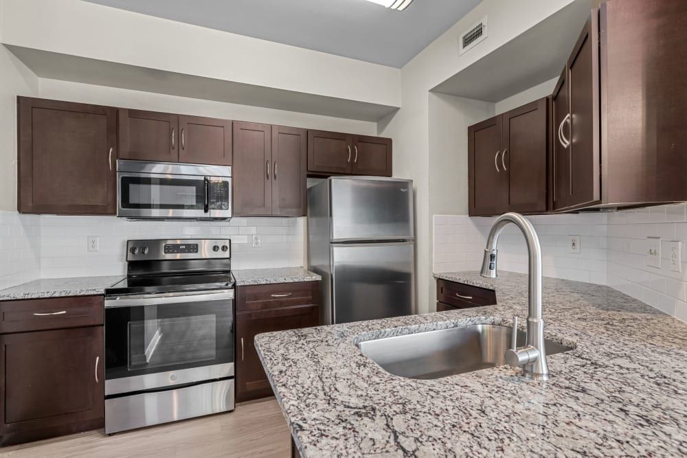Modern kitchen interior with dark brown wooden cabinets, stainless steel appliances including a microwave, oven, and refrigerator, and white subway tile backsplash. The kitchen features granite countertops with a deep sink and a high-arched faucet. No people are visible in the scene at Brooks on Preston in Plano, TX.