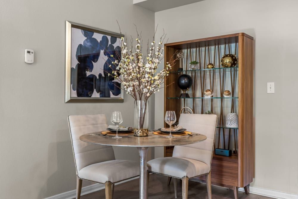 Elegant dining area with a round wooden table set for two, featuring cream upholstered chairs against a neutral wall. A modern, abstract painting in blue tones hangs on the left, and a sleek wooden shelving unit displaying decorative objects is on the right. Ambient light illuminates the room, casting a warm and inviting atmosphere at Brooks on Preston in Plano, TX.