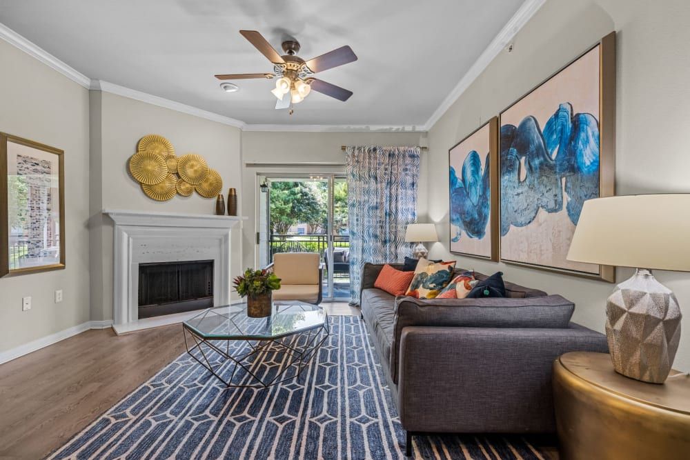 Elegantly decorated living room with a contemporary design. Features include hardwood floors, a white fireplace, and a ceiling fan. The room is furnished with a dark grey sectional sofa adorned with colorful pillows, a round glass coffee table on a patterned blue rug, a tan armchair, and oversized abstract wall art in blue tones. Gold decorative wall plates hang above the fireplace, and the room receives ample natural light from the sliding glass door leading to the outdoor area at Brooks on Preston in Plano, TX.