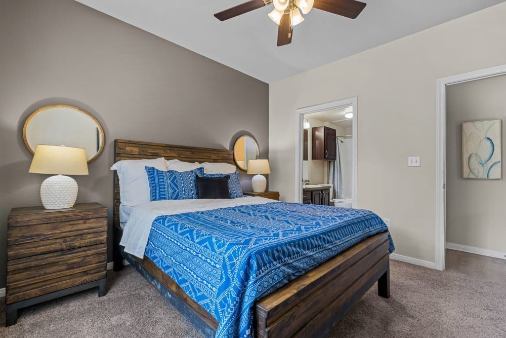 Elegantly decorated bedroom with a dark wood bed frame and a bright blue and white patterned bedding set. Two matching bedside tables each hold a white ceramic lamp with a round shade, flanked by round gold-framed mirrors on the wall above them. The room features beige walls and a plush carpet, with an overhead ceiling fan, and an open doorway leading to an ensuite bathroom in the background at Brooks on Preston in Plano, TX.