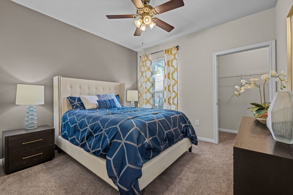 A neatly arranged bedroom with a queen-sized bed featuring a white tufted headboard and blue geometric-patterned bedding. Bedside tables with matching lamps flank the bed, with one showcasing a textured blue base. A brown dresser sits across the bed, adorned with a white orchid arrangement. The room has taupe walls, a light brown carpet, and a ceiling fan above. Bright sunlight filters through the window draped with patterned yellow curtains next to an open closet door at Brooks on Preston in Plano, TX.