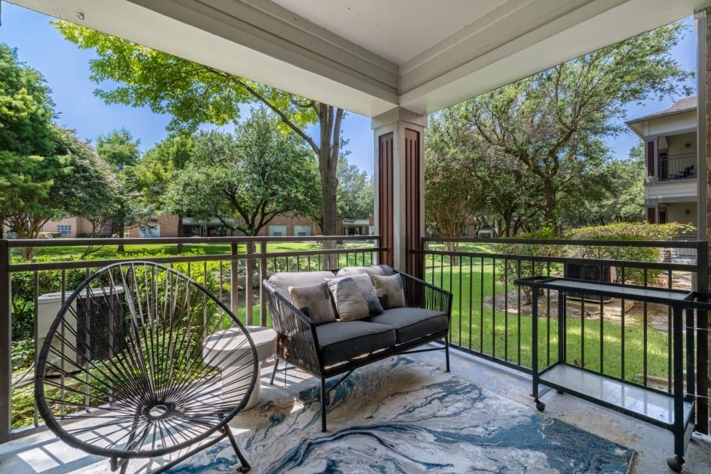 A cozy balcony space with a modern black circular chair and a cushioned lounge chair on a patterned area rug. The balcony railing overlooks a well-maintained communal garden with lush trees and green lawns, and the overhead roof provides shade on a sunny day at Brooks on Preston in Plano, TX.
