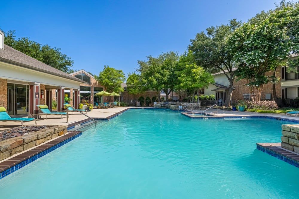 Tranquil outdoor pool area with clear blue water, surrounded by brick residential buildings and lounge chairs under shady trees on a sunny day at Brooks on Preston in Plano, TX.