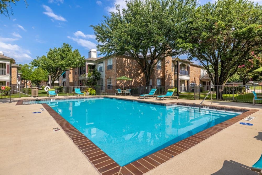 Inviting outdoor swimming pool with clear blue water surrounded by a well-maintained pool deck with lounge chairs, flanked by multi-story residential buildings and lush green trees under a bright blue sky at Brooks on Preston in Plano, TX.